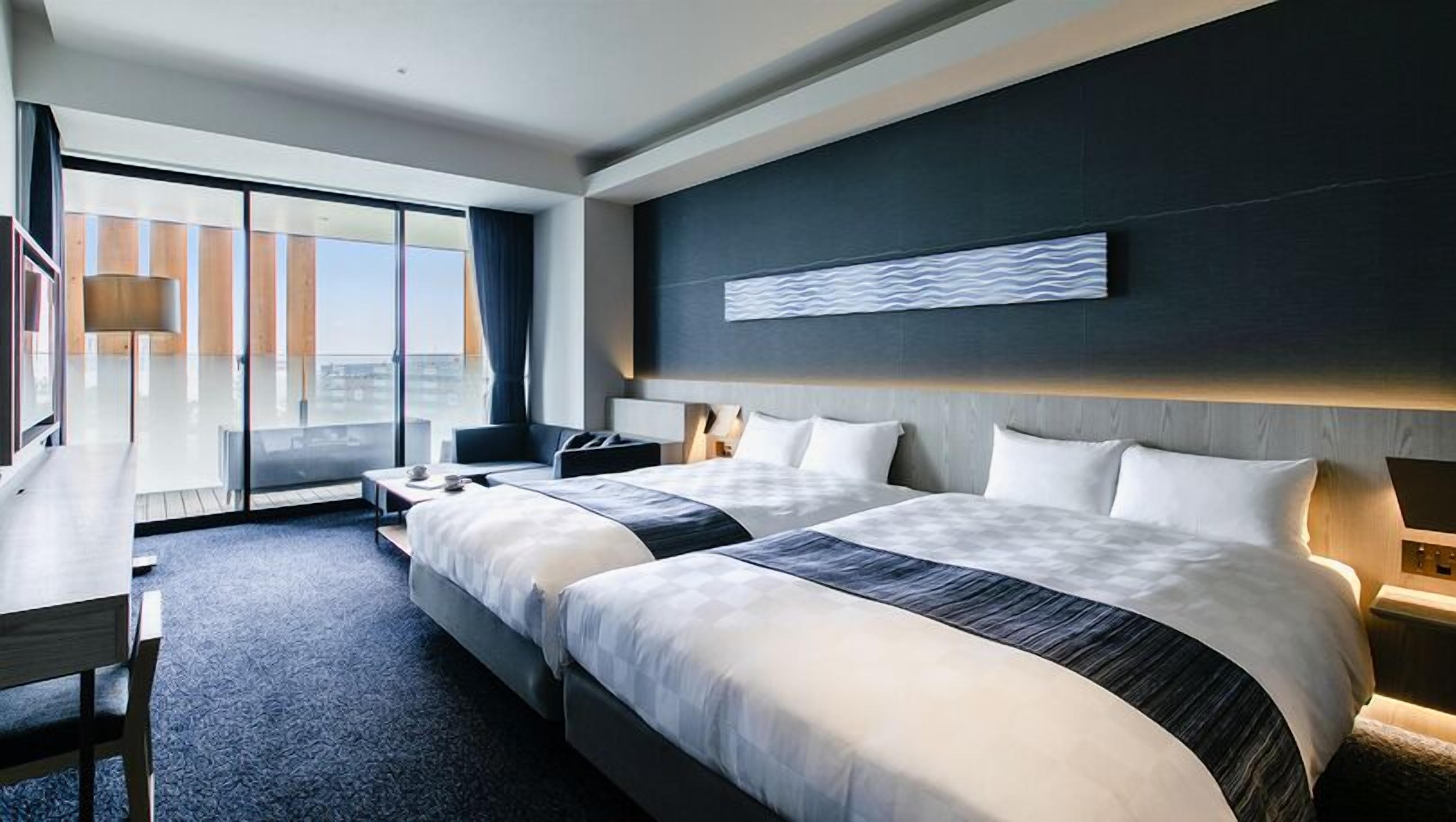 A modern hotel room with two beds, a desk, a chair, and a large window offering a city view.