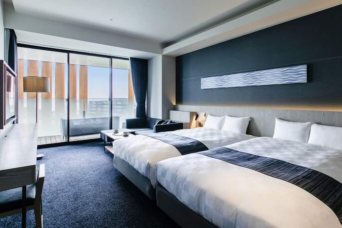 A modern hotel room with two beds, a desk, a chair, and a large window offering a city view.