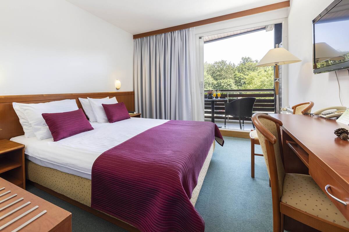 Une chambre d’hôtel avec un lit, un bureau, une chaise, une table de chevet et une fenêtre avec des rideaux.