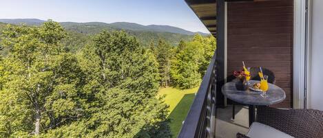 A balcony with a table set for two, overlooking a forested landscape with hills in the distance.