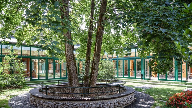 Um jardim circular de pedra e tijolo com grade de metal, rodeado por árvores e um edifício com portas de vidro e molduras verdes.