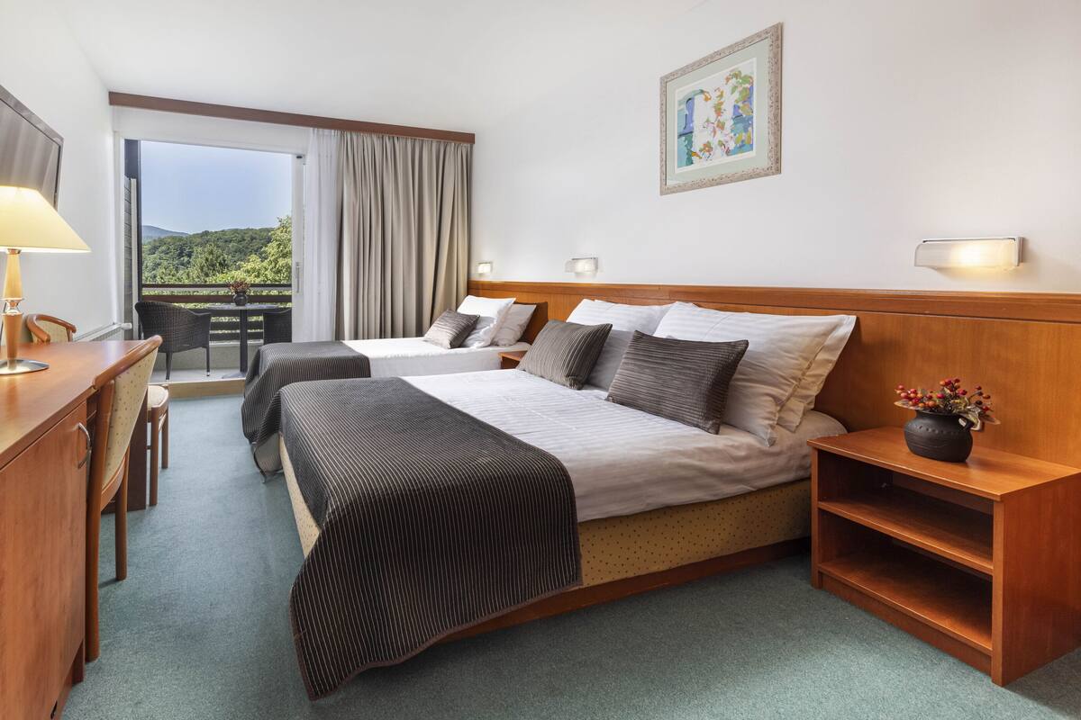 Une chambre d’hôtel avec deux lits, un bureau, une chaise, une petite table et un balcon donnant sur un espace verdoyant.