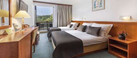 A hotel room with two beds, a desk, a chair, a small table, and a balcony with a view of greenery.