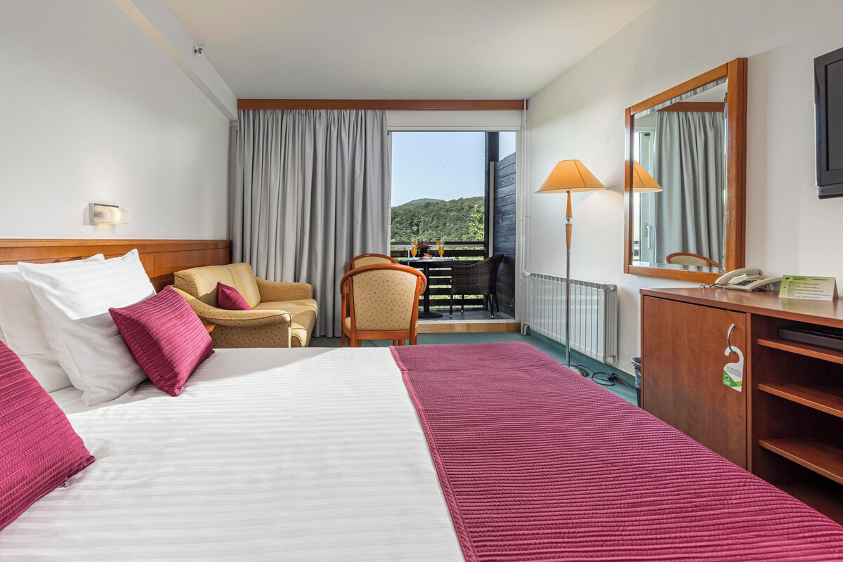 Habitación de hotel con cama, escritorio, silla, balcón con vistas y un espejo.