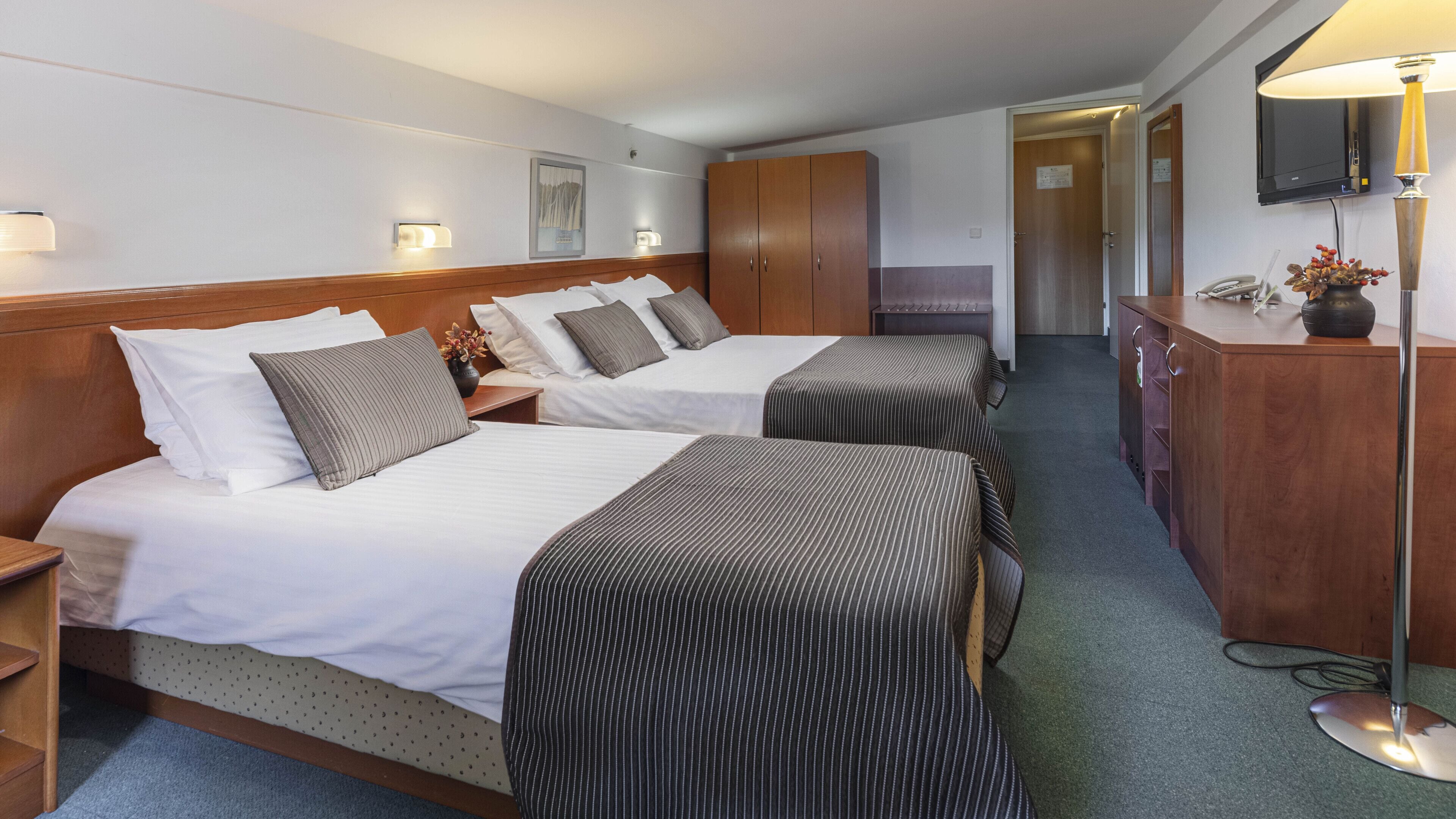 A hotel room with two beds, a wooden wardrobe, a desk with a lamp, and a television mounted on the wall.