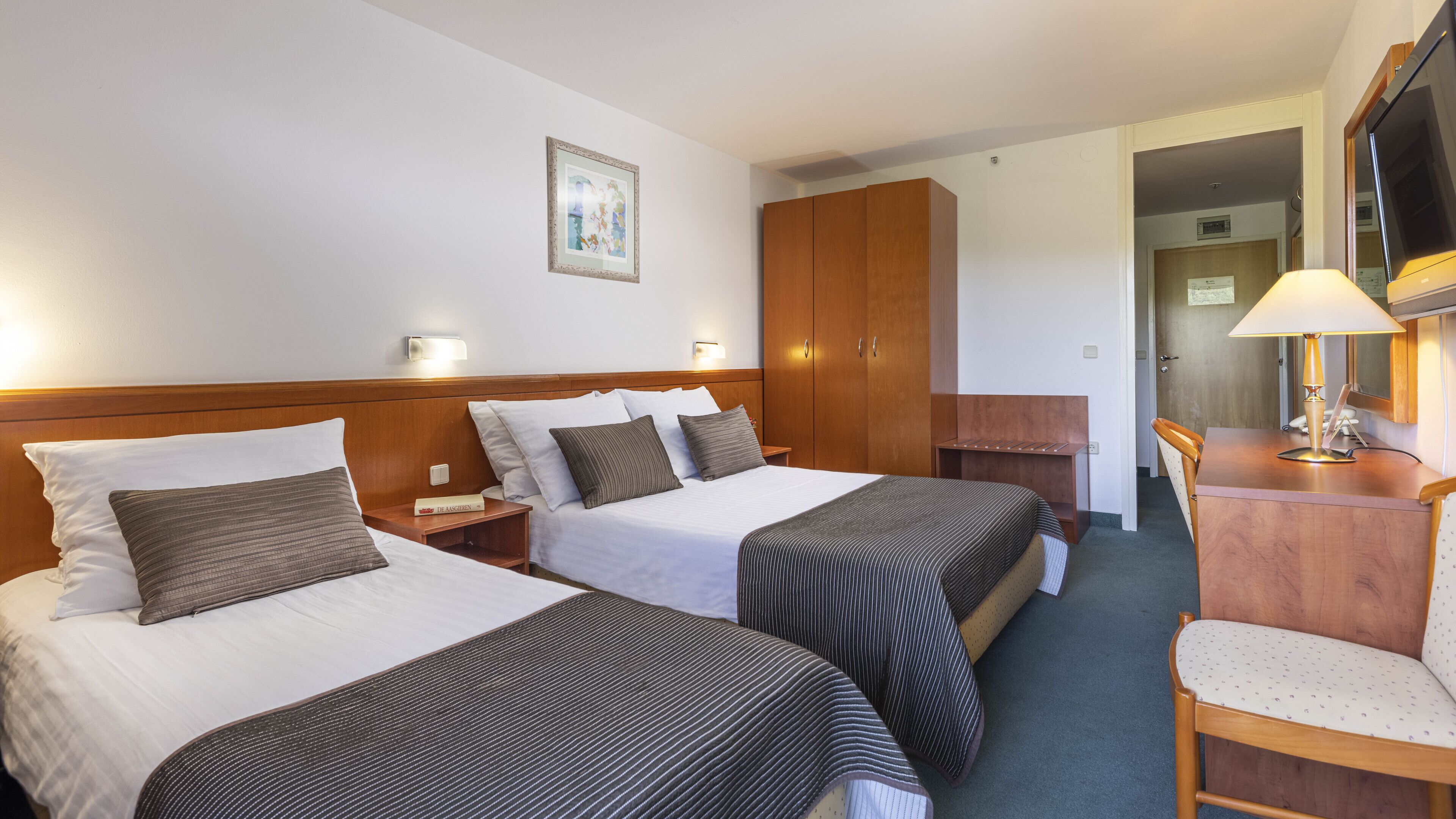 Ett hotellrum med två sängar, en garderob i trä, ett skrivbord med en lampa och en tv.
