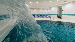 Piscina interna com efeito cascata, cadeiras de descanso azuis e parede revestida com faixa azul.