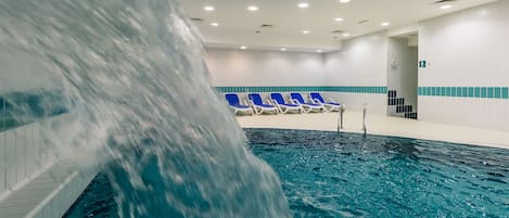 Une piscine intérieure agrémentée d’une cascade, de chaises longues bleues et d’un mur carrelé avec une bande bleue.