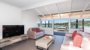 A modern living room with a flat-screen TV, two sofas, a coffee table, and large windows offering a view of a body of water and hills. - The Tide Watcher - Okiato Holiday Home (Okiato)