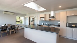 A modern kitchen with a large island, built-in appliances, and a dining area with a table and chairs. - The Tide Watcher - Okiato Holiday Home (Okiato)