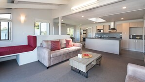 A modern living room with a sofa, a coffee table, and a kitchen area with cabinets and appliances. - The Tide Watcher - Okiato Holiday Home (Okiato)
