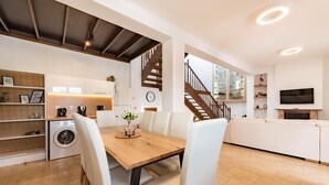 Una cucina moderna con un tavolo di legno, sedie bianche, una lavatrice e una scala che conduce a una zona living con televisore e camino.