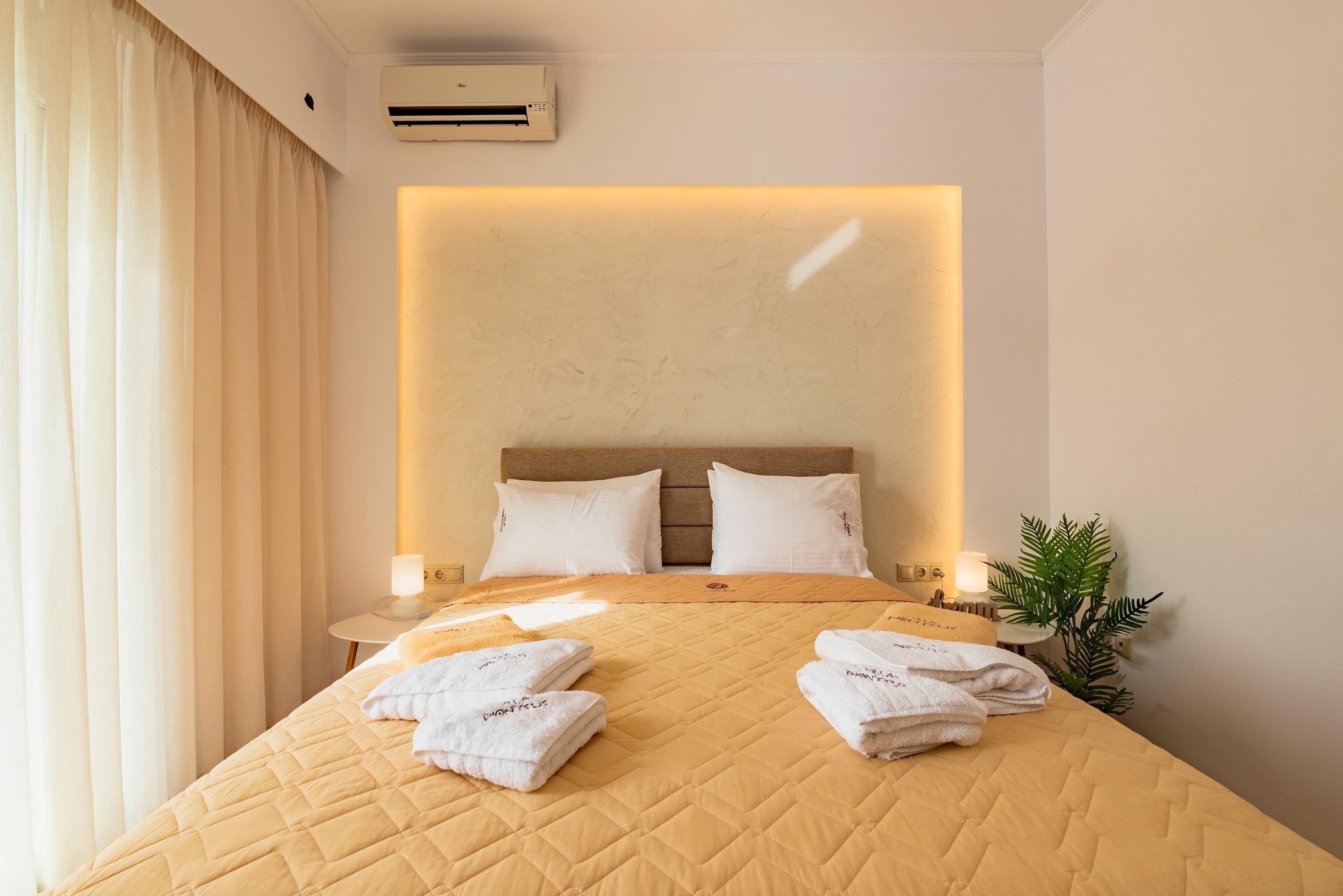 A hotel room with a bed, pillows, a bedside table with a lamp, a plant, and a wall-mounted air conditioner.