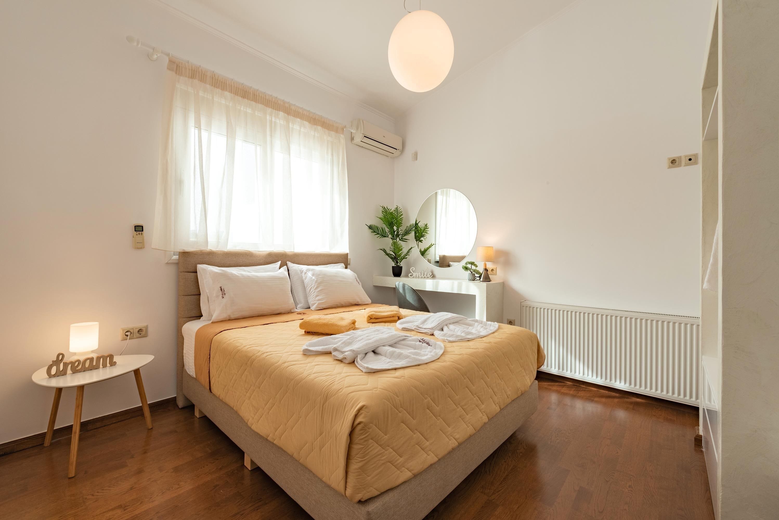 Un dormitorio con cama, mesita de noche, lámpara, equipo de aire acondicionado, ventana con cortinas, espejo y radiador.