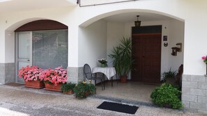 Una entrada de edificio con puerta, ventana con persianas, mesa con sillas y plantas en macetas.