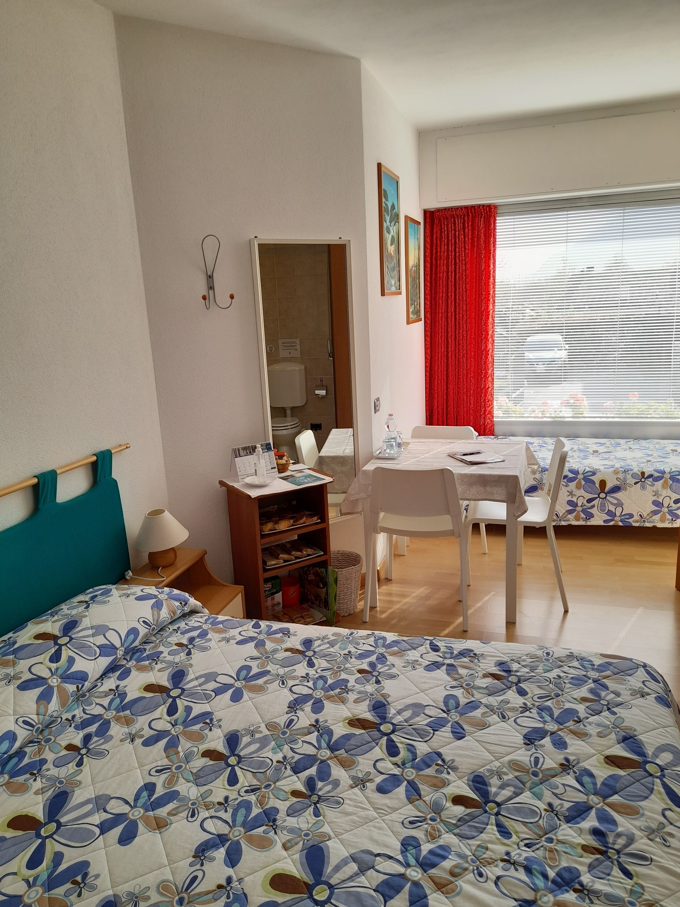 Una camera d'albergo con un letto, una scrivania, una sedia, un tavolino, uno specchio e una finestra con tende.