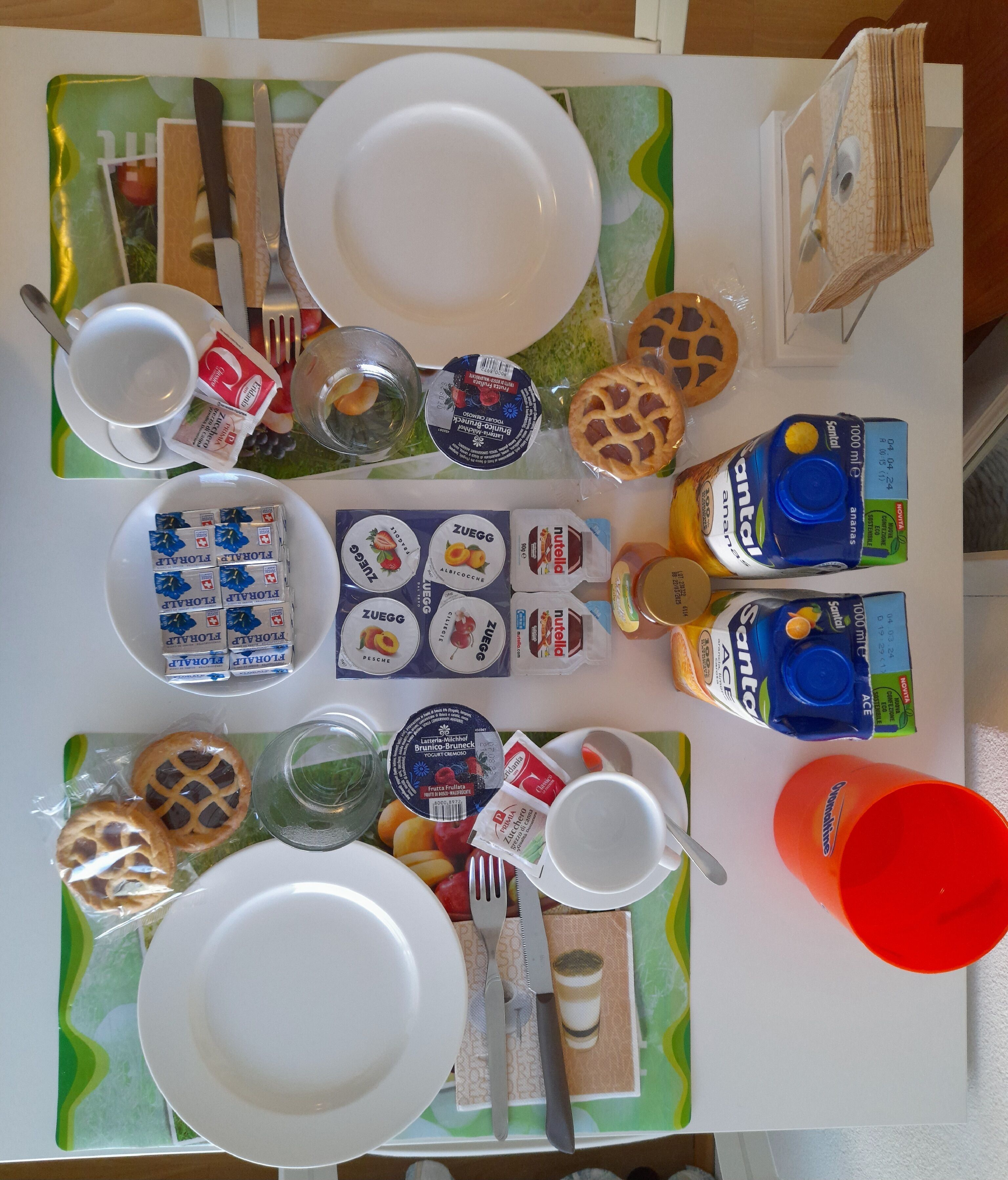 Ein Tisch, gedeckt mit Tellern, Besteck und verschiedenen Speisen, darunter verpackte Snacks und Getränke.