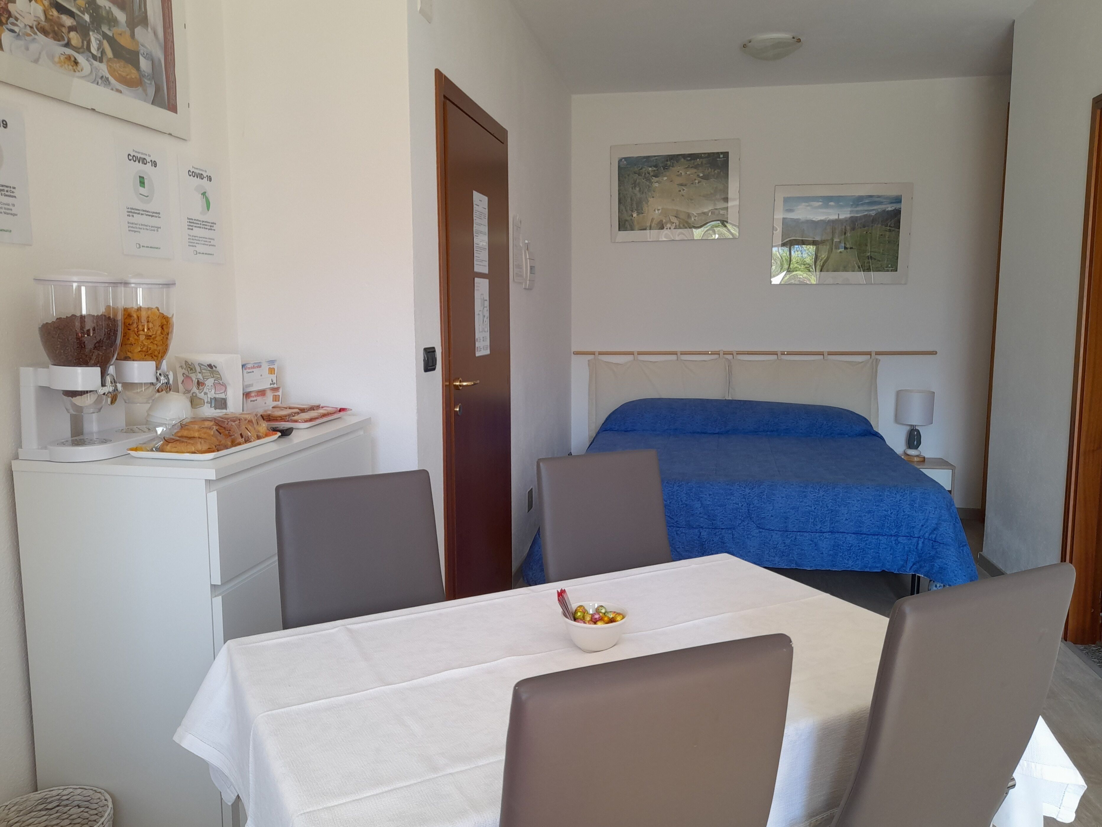 Una habitación de hotel con cama, una mesa y sillas, una barra de desayuno con comida y bebidas, y dos cuadros en la pared.