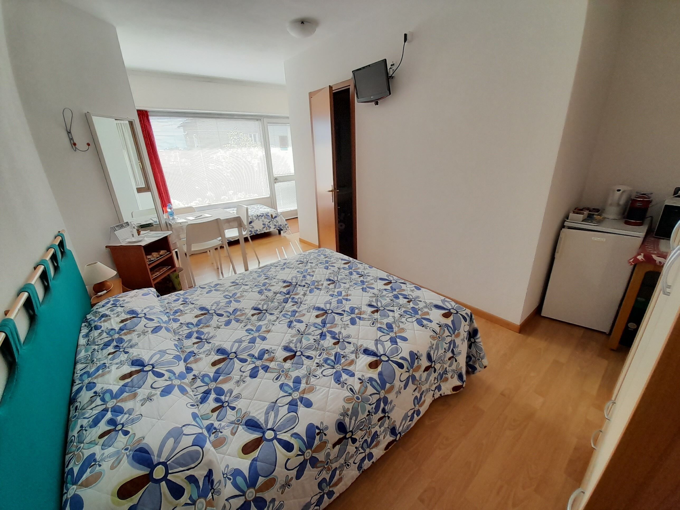 A bedroom with a bed, a desk, a chair, a small kitchenette, and a window with blinds.