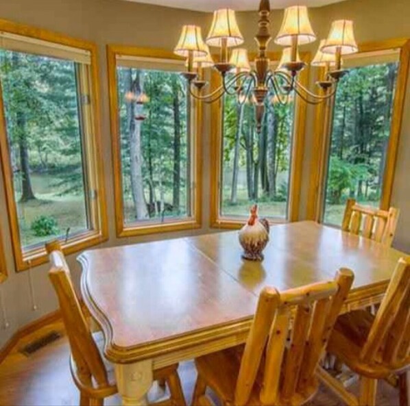 A dining area with a wooden table and chairs, surrounded by large windows with a forest view.