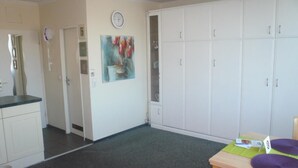 A room with a kitchenette, a dining table with a purple cushion, a clock on the wall, and a large white wardrobe. - App. Grimme 09 O, Germany (Sylt)
