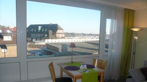A room with a dining table, chairs, a lamp, and a view of buildings and a dock through large windows. - App. Grimme 09 O, Germany (Sylt)