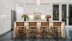 A modern kitchen with a central island, three wicker chairs, a chandelier, and a large window.