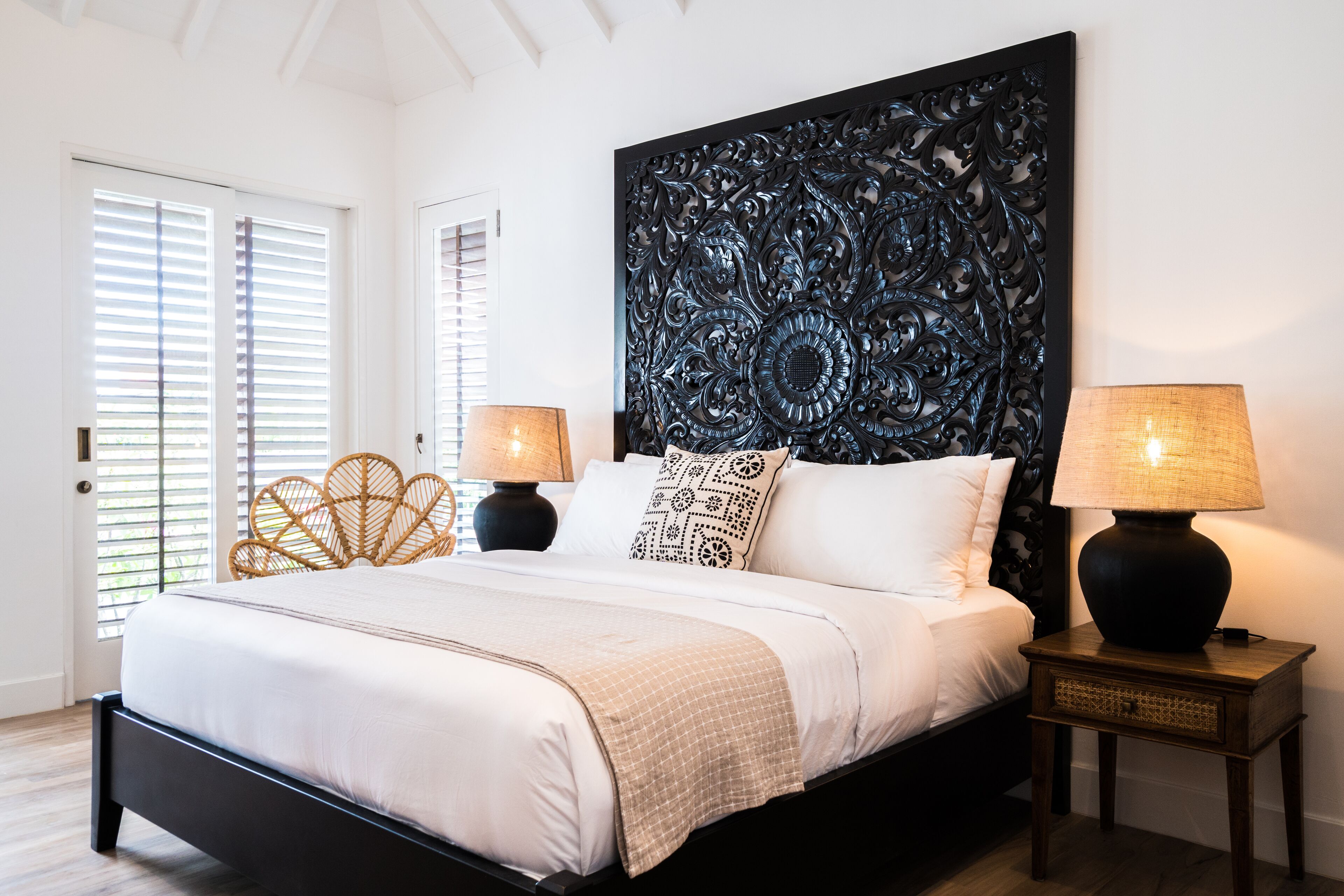 A bedroom with a large bed, two bedside tables with lamps, a decorative headboard, and a door with blinds.