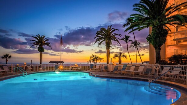 A swimming pool with lounge chairs and palm trees, surrounded by a fence, during sunset.