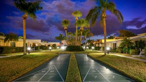 A nighttime view of a courtyard with a shuffleboard court, palm trees, and illuminated buildings.