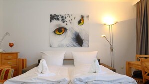 A hotel room with a bed, two pillows, a bedside table with a lamp, a chair, and a painting of an owl's face on the wall.