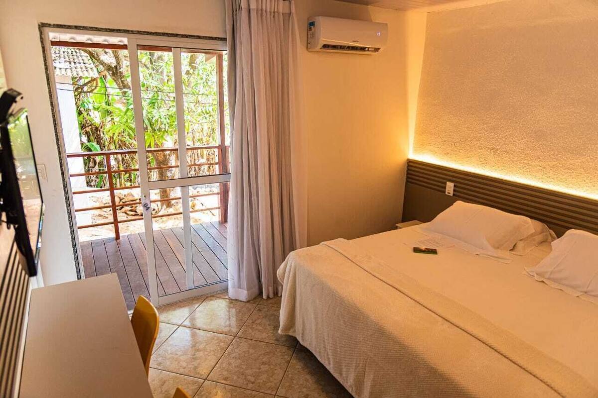 A hotel room with a bed, a desk with a chair, a balcony with a view of greenery, and an air conditioning unit.