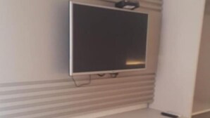 Quarto compacto com televisão fixada na parede, um refrigerador branco pequeno e uma cadeira amarela.