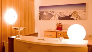 Una habitación de hotel con pared de paneles de madera, una gran fotografía enmarcada de un paisaje montañoso, un espejo redondo en la tocador, y un pequeño aparador de madera con un bolso rojo y un jarrón blanco.