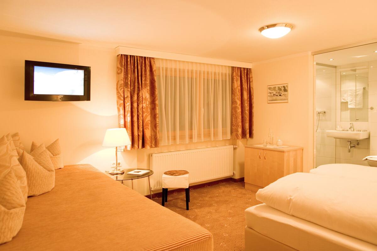 Habitación de hotel con cama, una mesita con lámpara, ventana con cortinas, radiador, baño con lavamanos y espejo, y un cuadro en la pared.