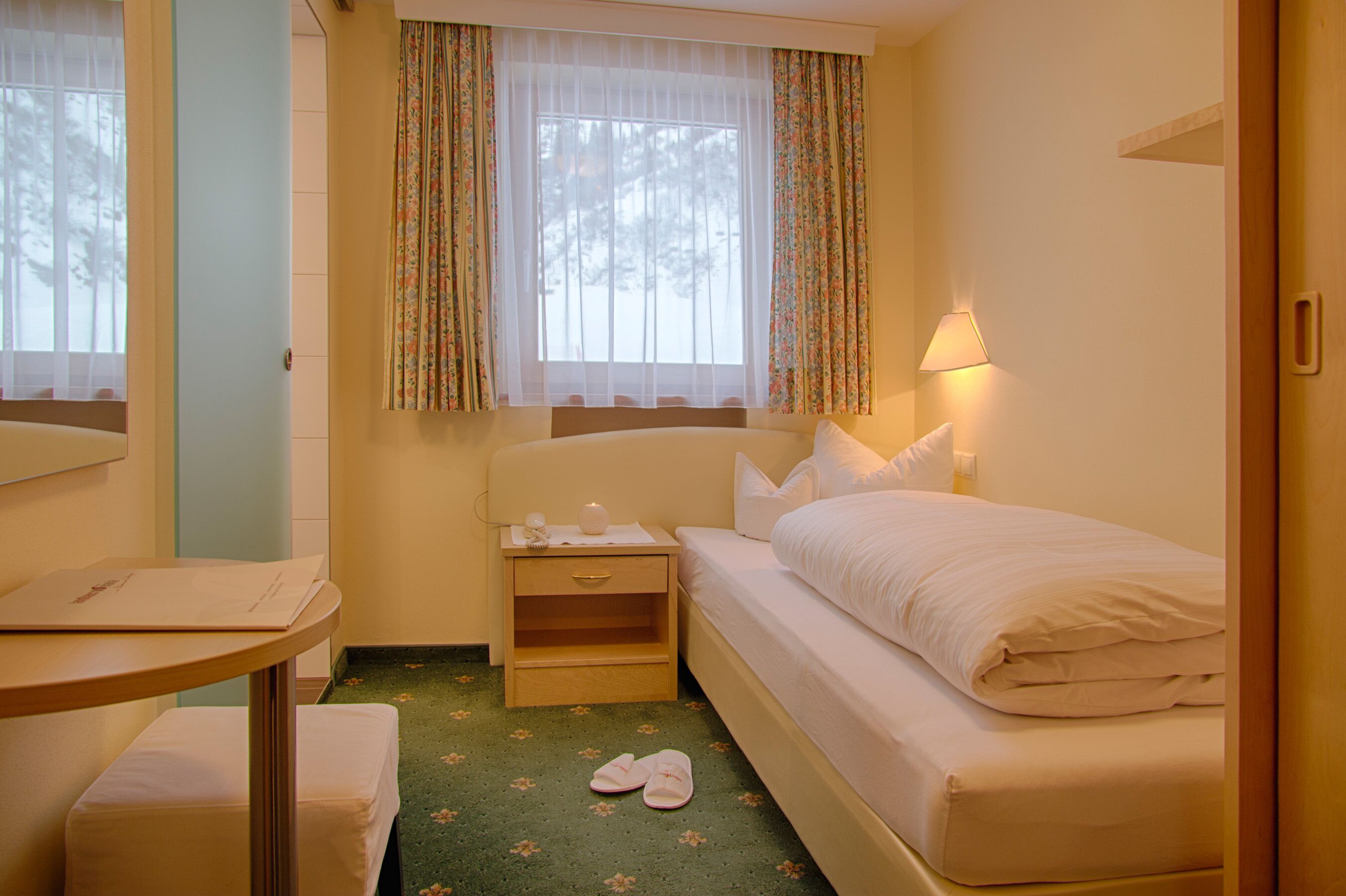 A small, cozy hotel room with a single bed, a bedside table, a nightstand, a lamp, and a window with floral curtains.