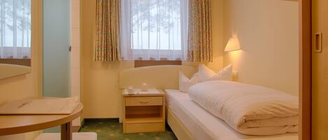 A small, cozy hotel room with a single bed, a bedside table, a nightstand, a lamp, and a window with floral curtains.