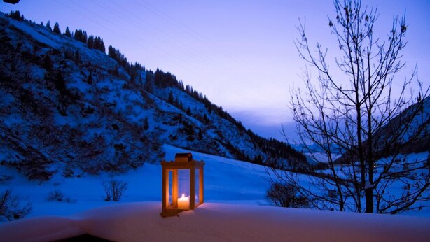 Paisaje nevado con un farol iluminado sobre una plataforma, rodeado de árboles y una colina al fondo.