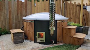 Una estufa de leña con chimenea, rodeada por una valla de madera y un patio con un banco y una mesita.