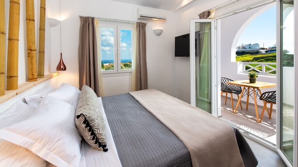 A bedroom with a bed, a window with curtains, a table with chairs, and a view of the landscape. - Abasa Suites (Santorini)