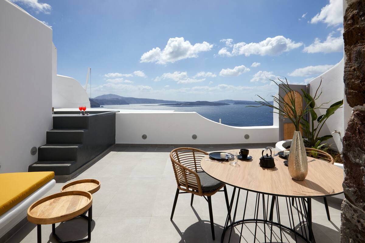 A modern outdoor dining area with a view of the sea, a wooden table, chairs, and a vase.
