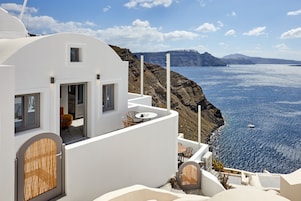 Un edificio blanco con balcón que da a la costa, con una mesa y sillas, y vistas al mar y montañas a lo lejos.