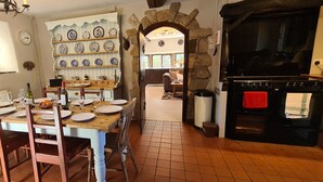 A dining area with a wooden table set for four, surrounded by chairs. A stone wall with shelves of plates is visible in the background. - Greave Farm Todmorden Tranquil Cottage Retreat (Todmorden)