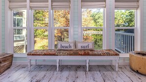 A bench with two pillows labeled "beach" and "surf" in front of a set of windows with blinds, overlooking a garden and a wooden deck. - 8852 Bay Pine 3 Bedroom Home by RedAwning (Miramar Beach)