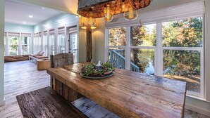 A spacious dining area with a wooden table, rustic pendant lighting, and a view of a deck and trees through large windows. - 8852 Bay Pine 3 Bedroom Home by RedAwning (Miramar Beach)