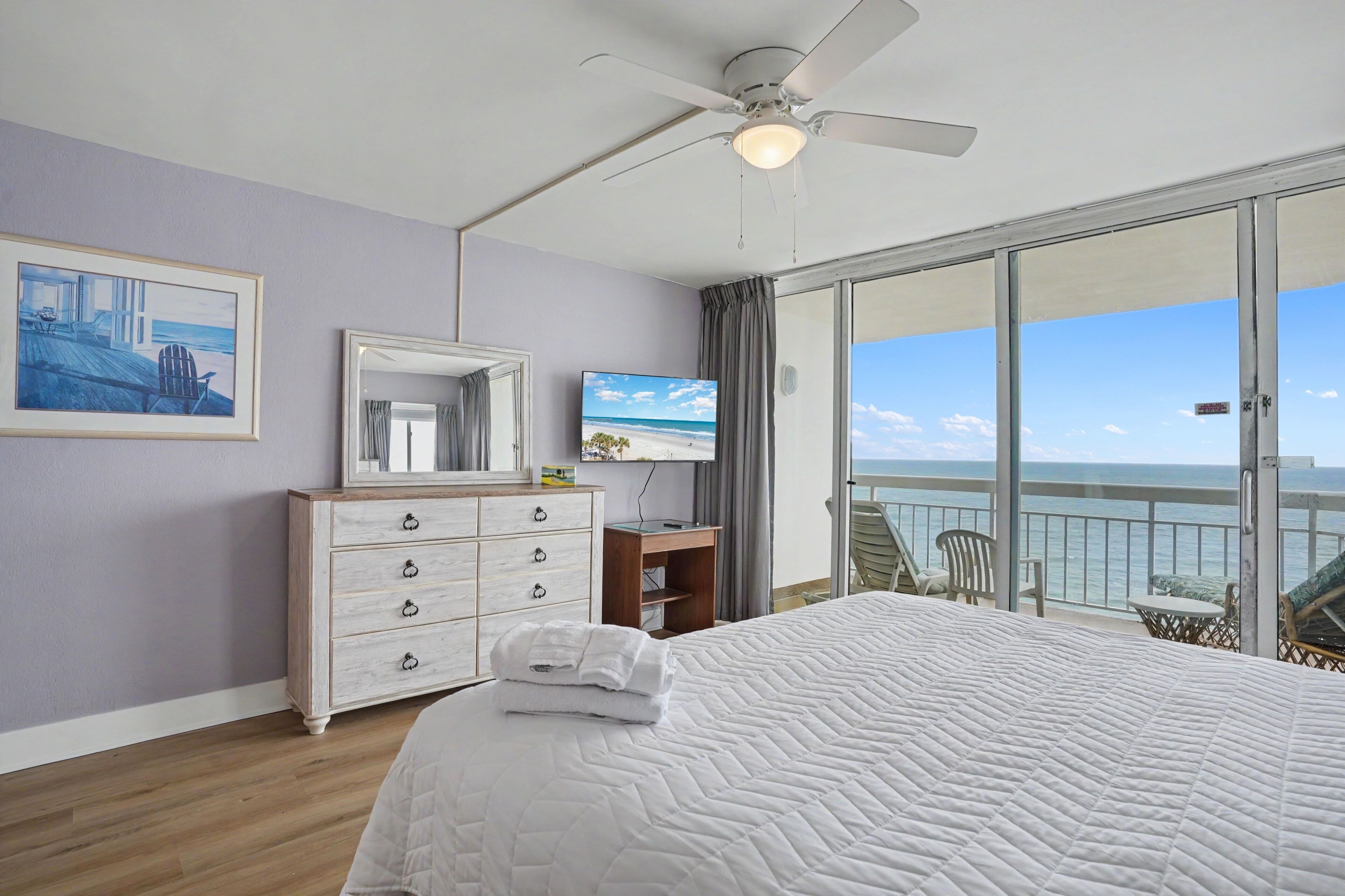 A bedroom with a bed, a dresser, a television, and a balcony with a view of the ocean.