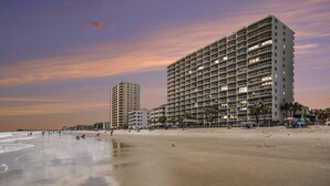 Un hotel frente a la playa con varios pisos, una playa de arena y un muelle al fondo.