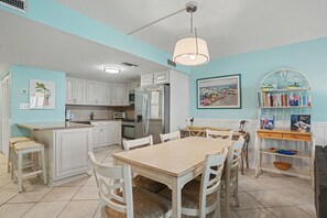 A kitchen with white cabinets, a dining table, chairs, a refrigerator, and a bookshelf.