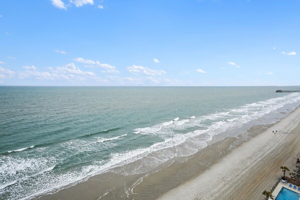A beach with a pier, clear skies, and a pool area with lounge chairs.
