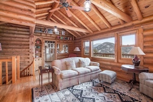 Interior de una cabaña de madera con techo de madera, un sofá, alfombra estampada y vistas a montañas a través de amplias ventanas.